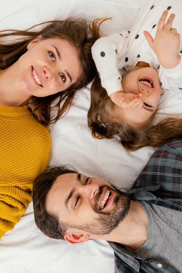 Fotografía de una pareja tumbada en la cama con su hija, sonriendo, plano cenital.