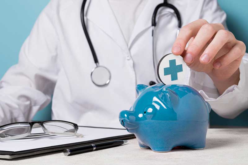 Fotografía de una mano y una hucha de cerdito azul. De fondo un profesional médico. Representación del ahorro en el seguro médico y de salud.