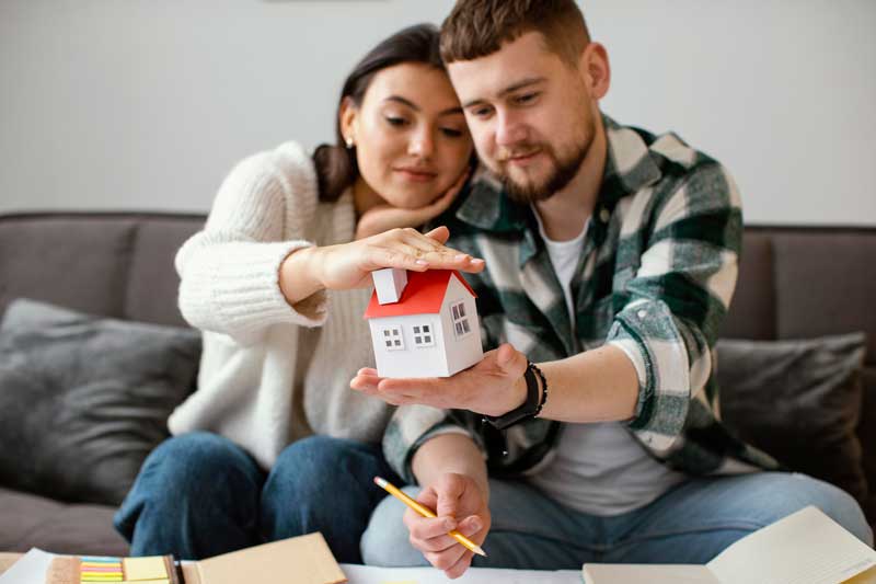 Pareja sentada en el sofá con una casa de madera en miniatura, ideando su hogar ideal.